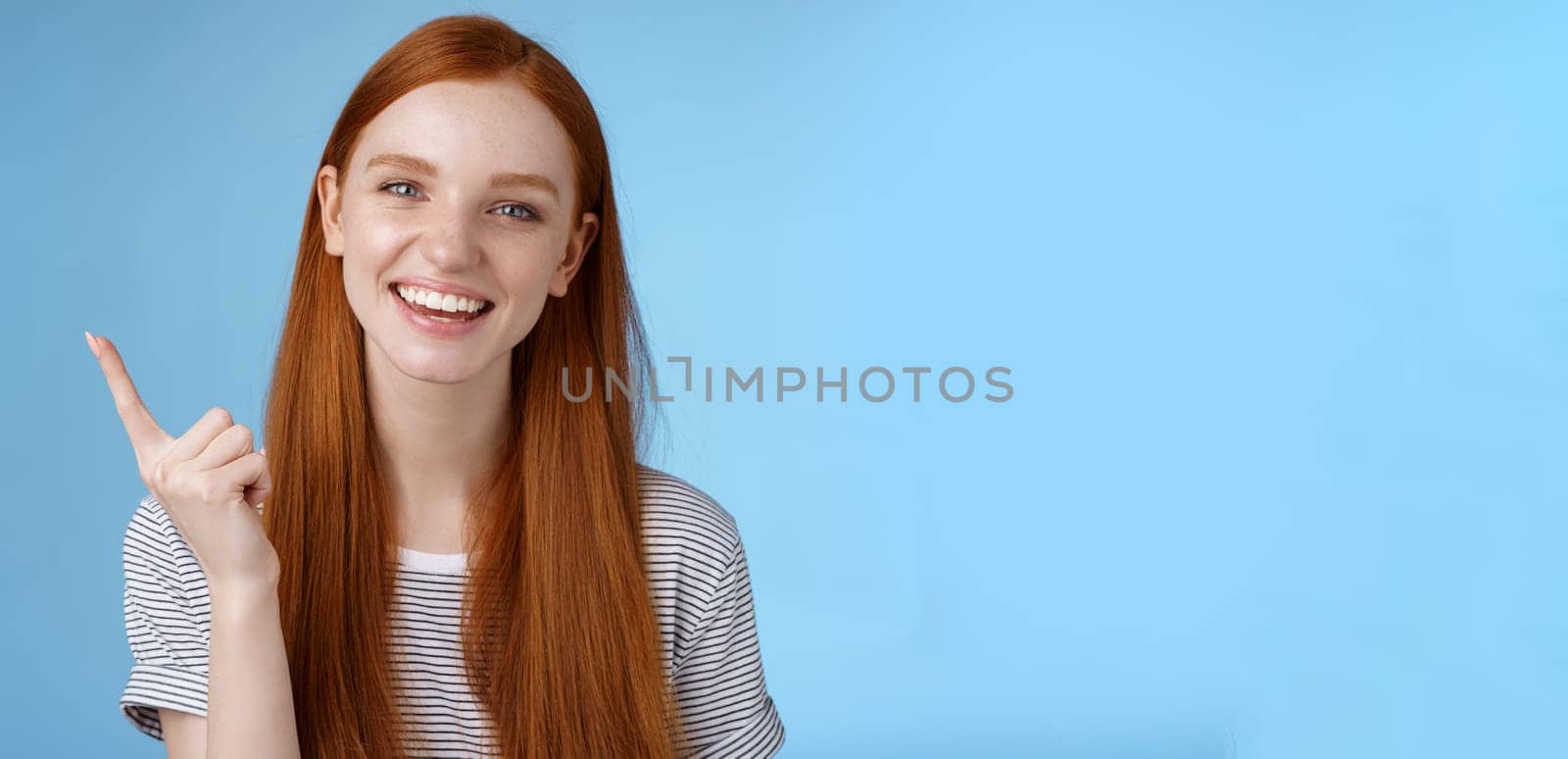 Happy charismatic redhead laughing young girl having fun looking carefree talking discussing new product sale pointing up index finger showing copy space advertisement, standing blue background.