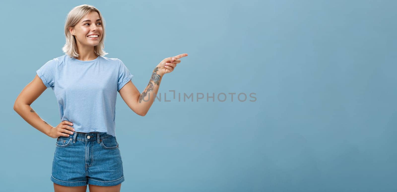 Bossy confident female manager with tattoo on arm holding hand on waist pointing and gazing right with pleased relaxed look giving directions to employees posing over blue background by Benzoix