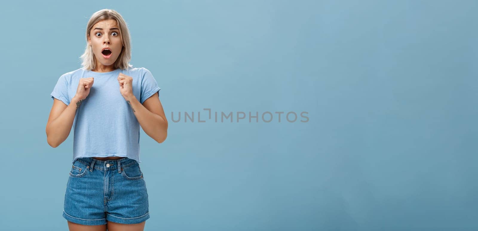 Studio shot of stunned shocked girl standing in stupor with dropped jaw and frightened look clenchign fists near breast from fear standing astonished over blue background by Benzoix