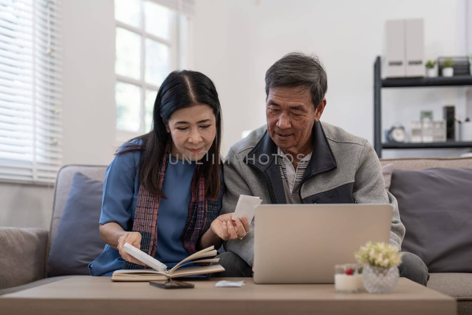Asian senior couple checking and calculate financial bill together on sofa involved in financial paperwork, paying tax online using laptop at living room home by nateemee