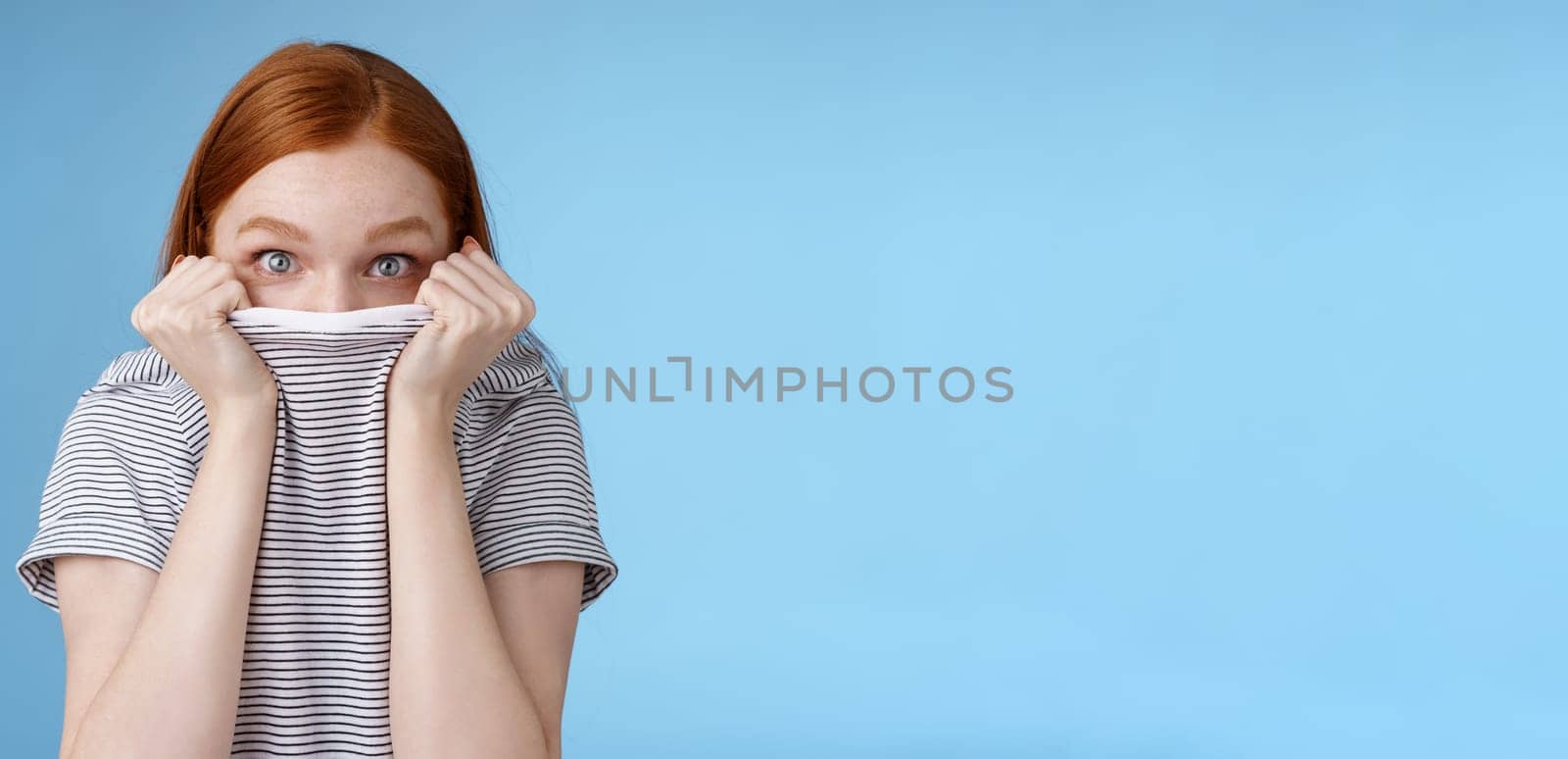 You cannot see me. Silly cute young teenage redhead girl hiding face pulling t-shirt head peeking look excited surprised camera hold collar standing funny under disguise, blue background.