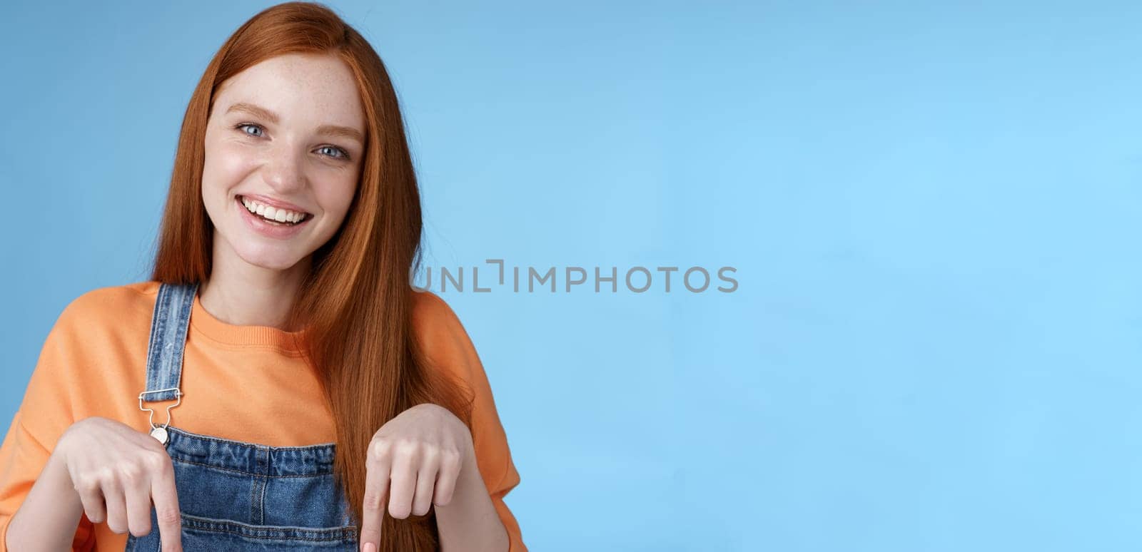 Friendly happy redhead girl smiling lively pretty grin pointing down index fingers offering good offer recommend use service standing blue background discuss interesting product, blue background.