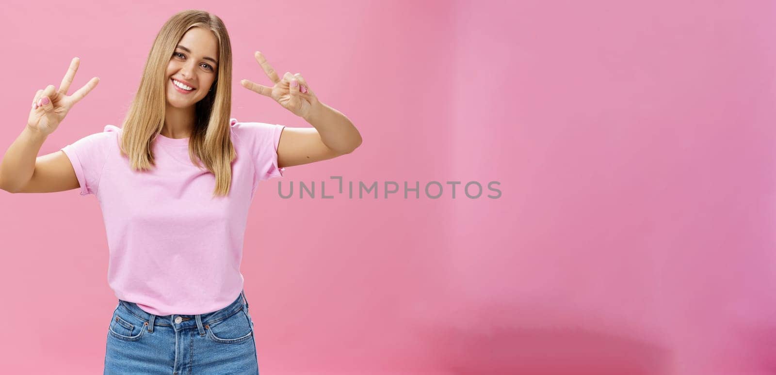 Cute chubby carefree young woman with short fair hair tilting head showing peace gestures with happy friendly smile having fun spending time amused and joyful against pink background. Copy space