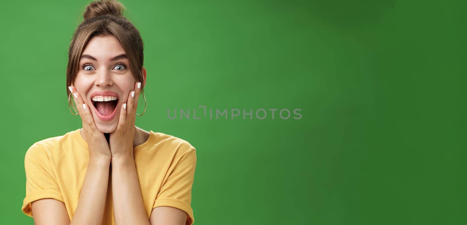 Portrait of happy delighted and surprised young feminine girl in yellow t-shirt pressing hands to cheeks from amazement and joy smiling broadly reacting to astonishing news over green background. Emotions concept