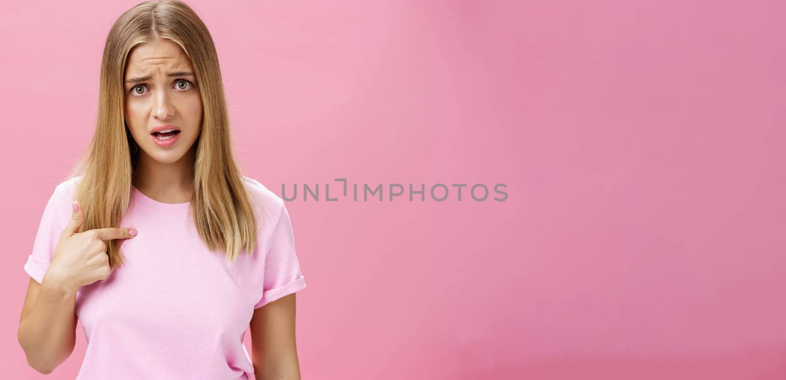 Insulted girl pointing at herself with displeased pissed and questioned expression asking question being shocked she picked or accused in something terrible and disrespectful posing against pink wall. Lifestyle.