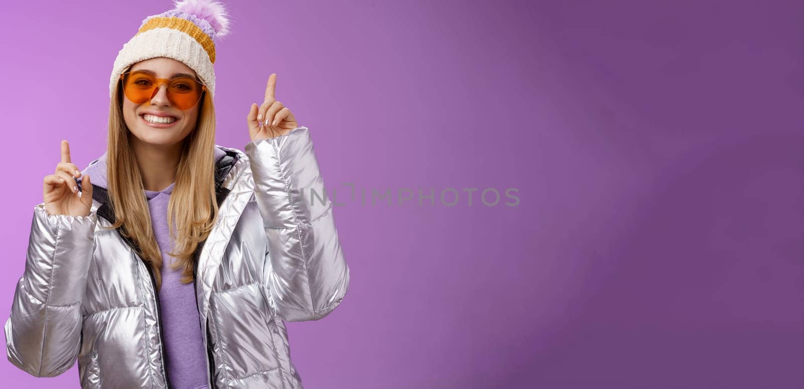 Lifestyle. Joyful energized entertained cute blond woman having fun enjoy vacation snowy mountain trip wearing sunglasses silver jacket winter hat dancing pointing up amused standing purple background.