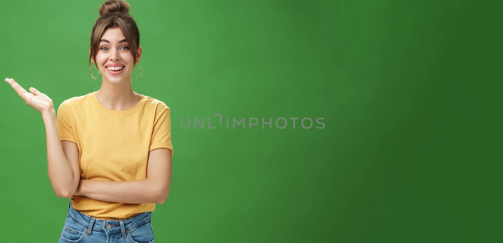 Wondered and carefree young friendly woman with combed hair cute gapped teeth in yellow t-shirt raising one hand in surprise reacting to amazing news smiling broadly at camera over green wall. Lifestyle.
