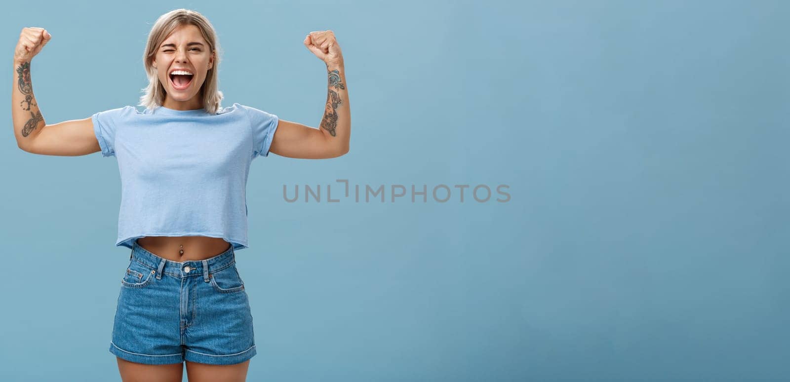 Strong women stand up and fight for rights. Portrait of happy entertained and cool young female athletic blonde with tattoos winking and smiling showing muscles and biceps over blue wall.