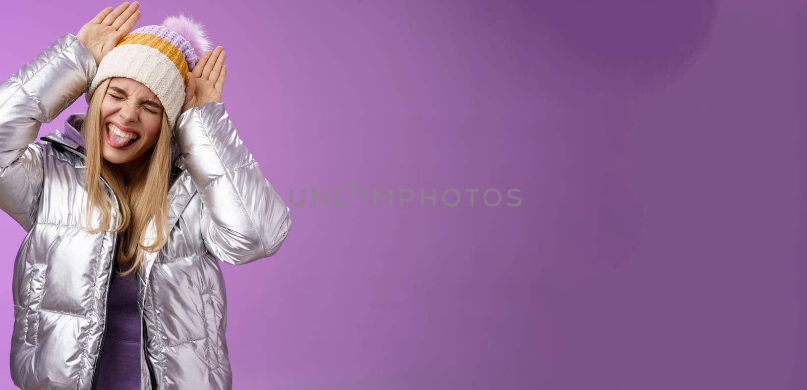 Carefree funny amused fair-haired caucasian girl 25s having fun playing around mimicking bunny close eyes show tongue excited make animal ears palms on head standing purple background joyful by Benzoix