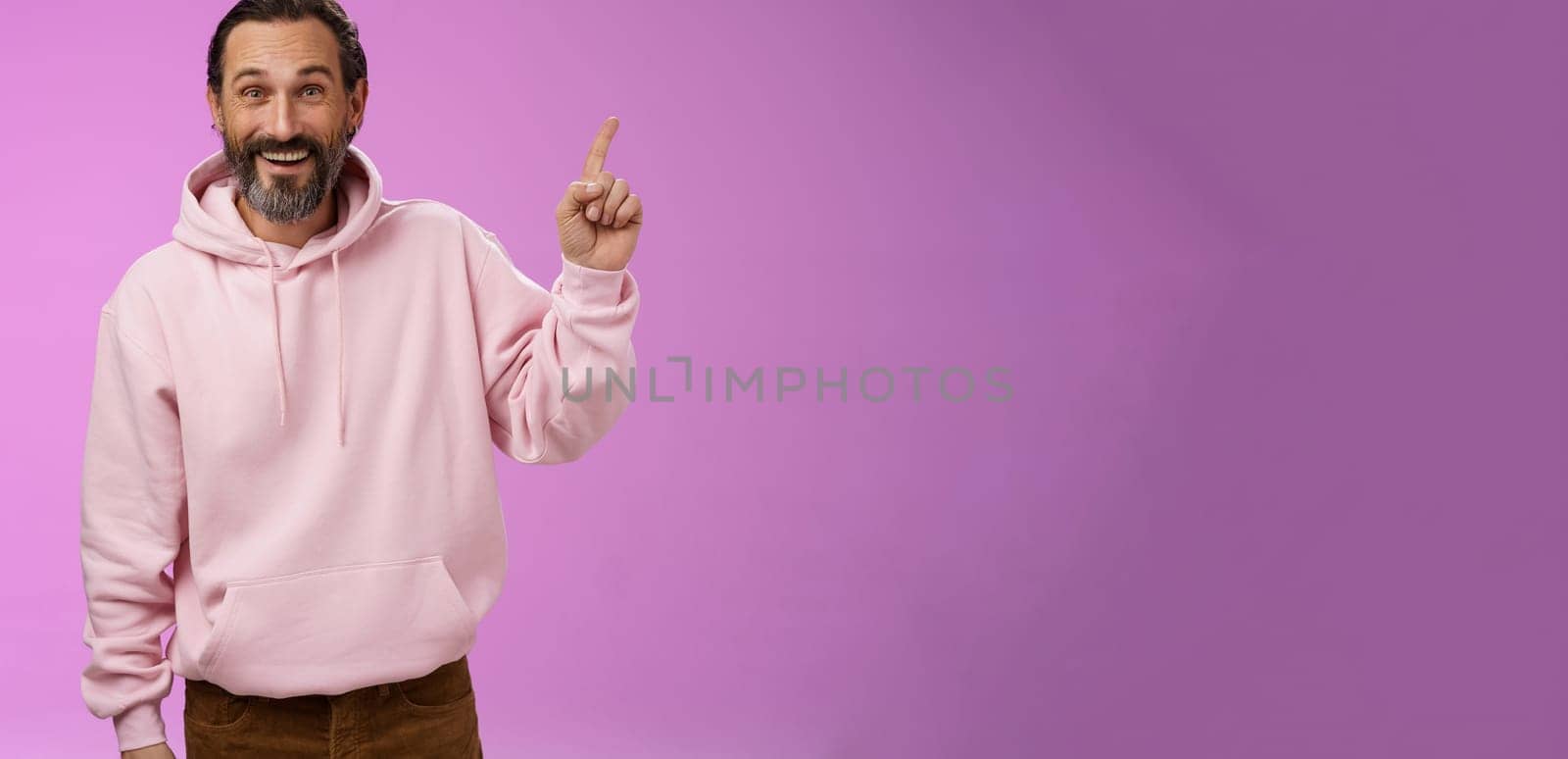 Handsome excited mature 40s bearded man wrinkles grey hair laughing happily acting immature having fun amusing vacation trip pointing up astonished smiling impressed surprised, purple background.