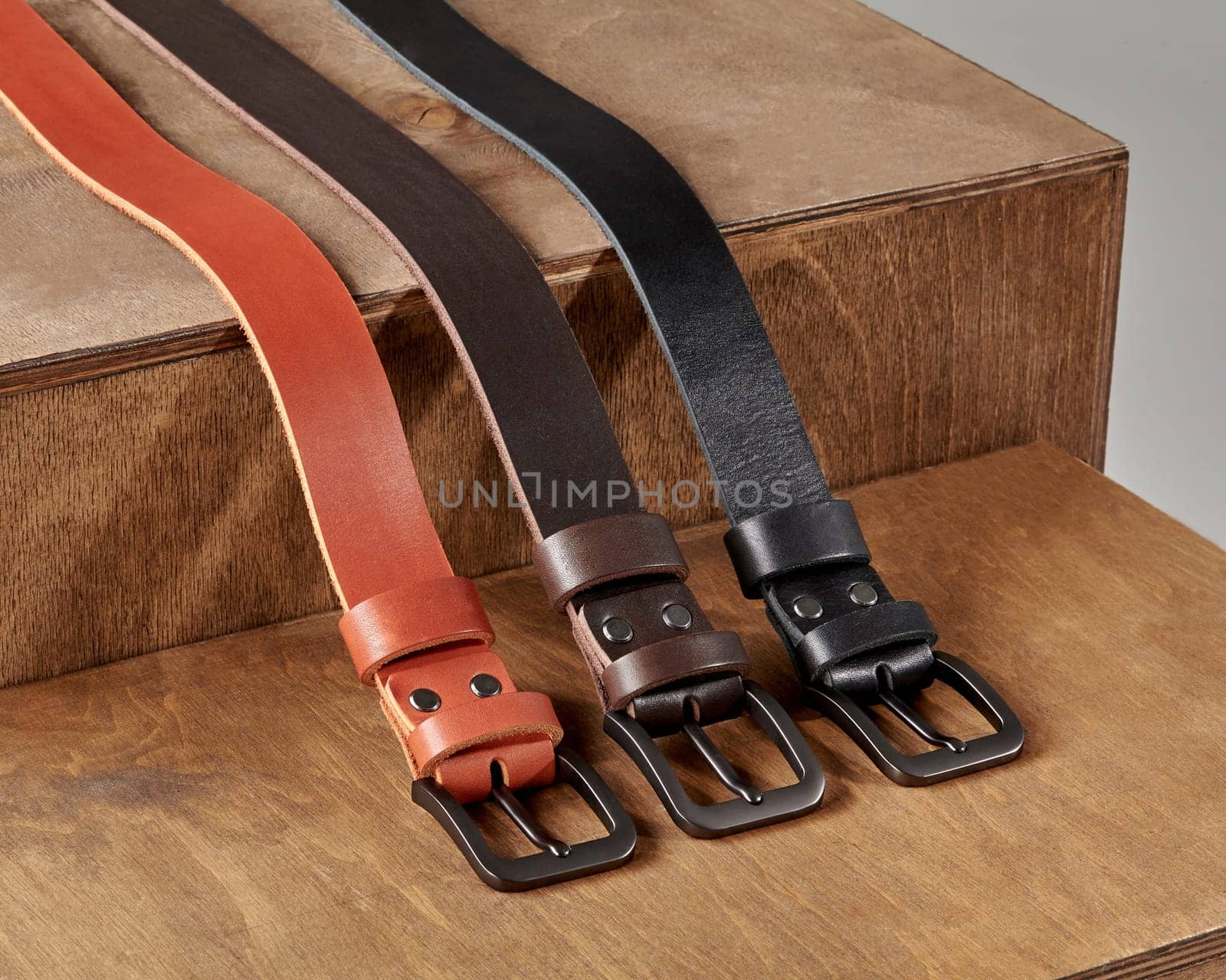 Three genuine leather belts of different colors with metal buckles and DAD embossing on loop displayed on wooden stand. Stylish personalized handcrafted mens accessories