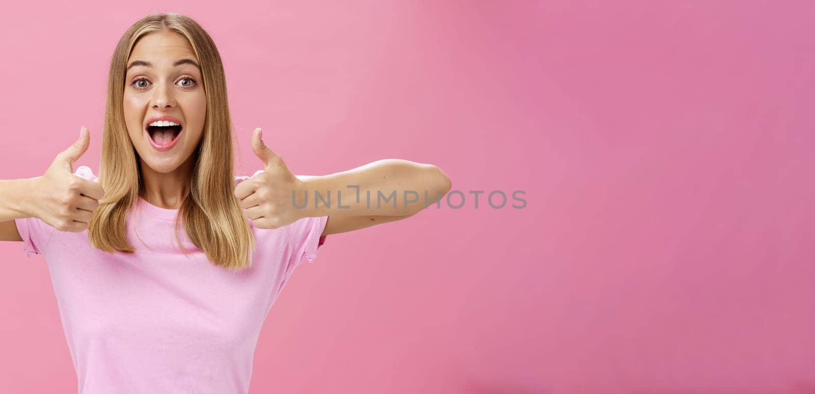 Supportive girlfriend cheering for favorite player showing thumbs up with happy positive smile giving approval and showing she likes perfect idea posing amused and enthusiastic against pink background. Lifestyle.