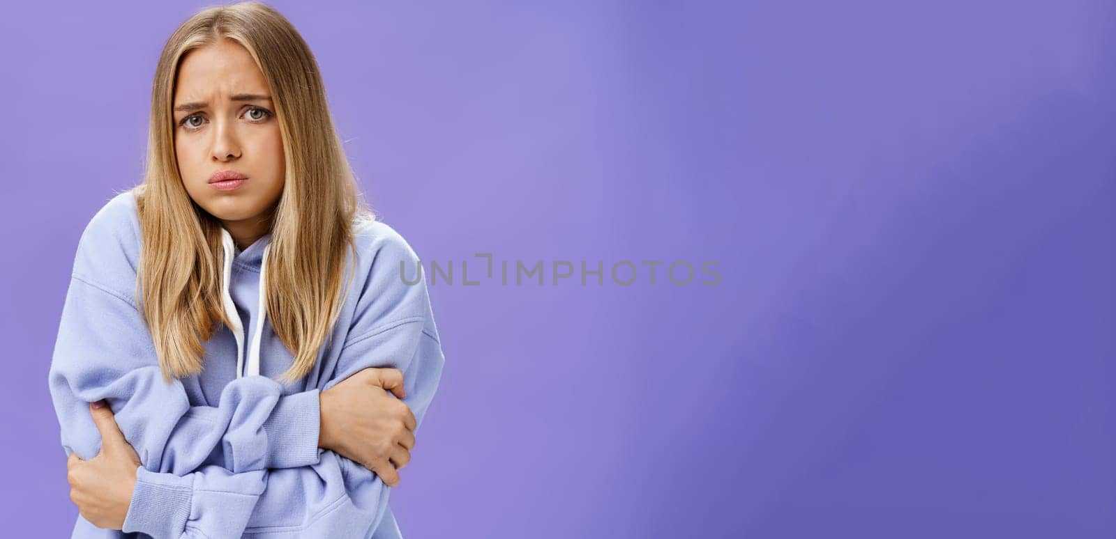 Girl freezing cold during chilly autumn evening trembling hugging herself with hands crossed against chest sulking, frowning wearing hoodie walking on windy and rainy weather posing over purple wall. Clothes, temprature and forecast concept