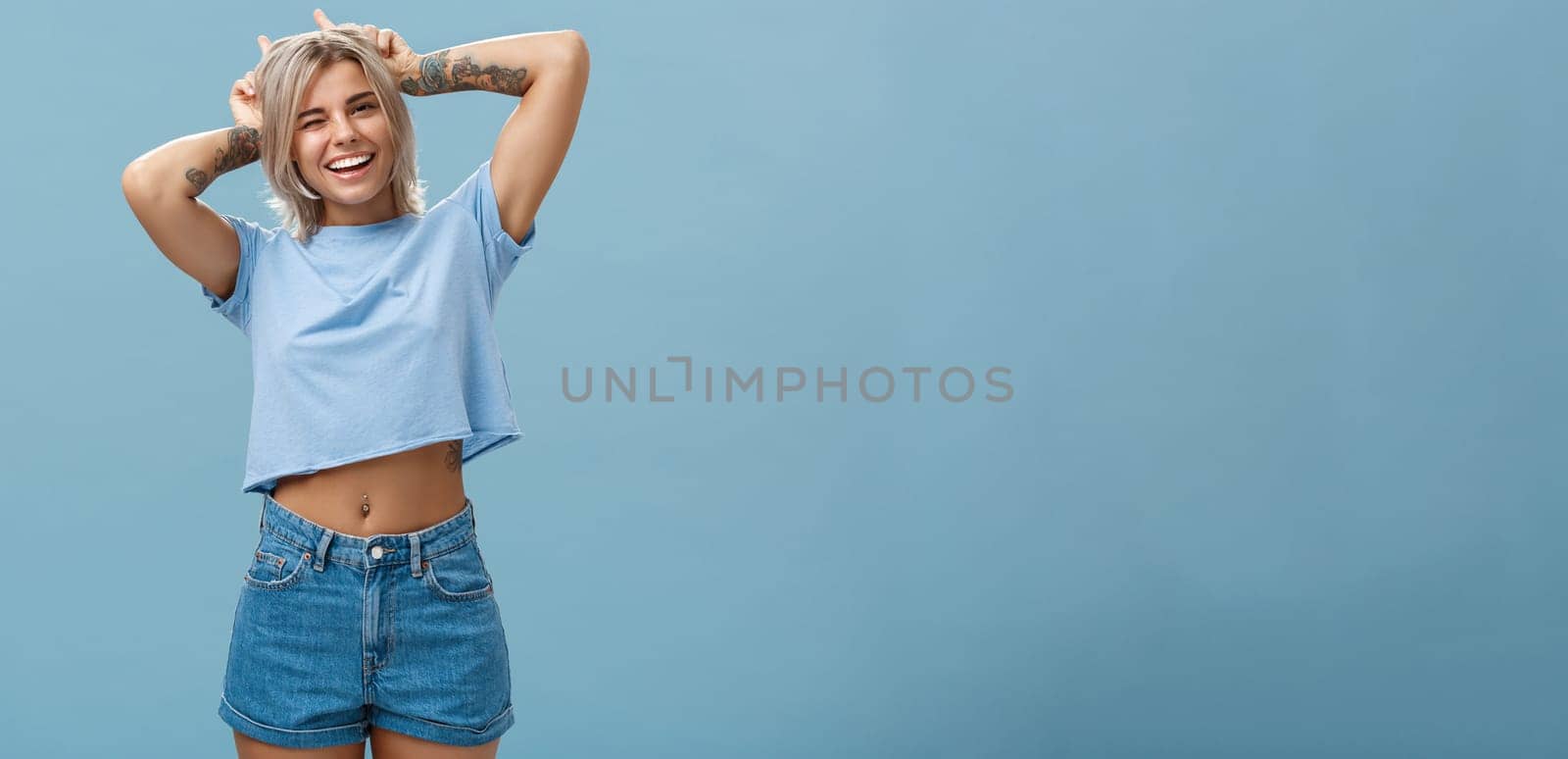 Studio shot of cute attractive and joyful stylish blond woman with tattoos on arms winking and smiling broadly holding index fingers on head making horns standing in summer outfit over blue wall by Benzoix