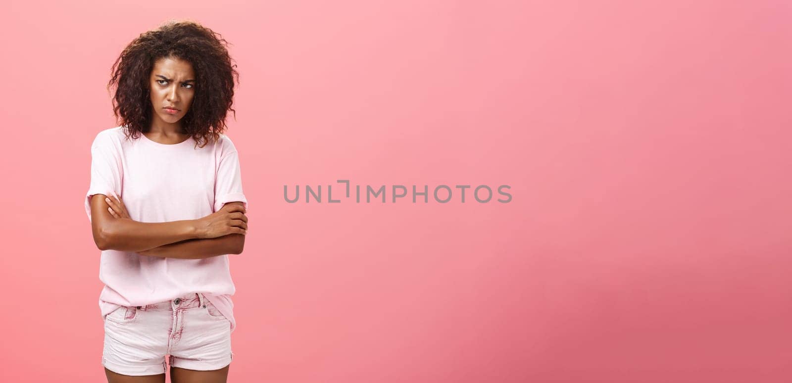 Portrait of offended gloomy sad african american female friend crossing arms on chest in protection gesture frowning looking from under forehead with insult looking envious and angry over pink wall by Benzoix