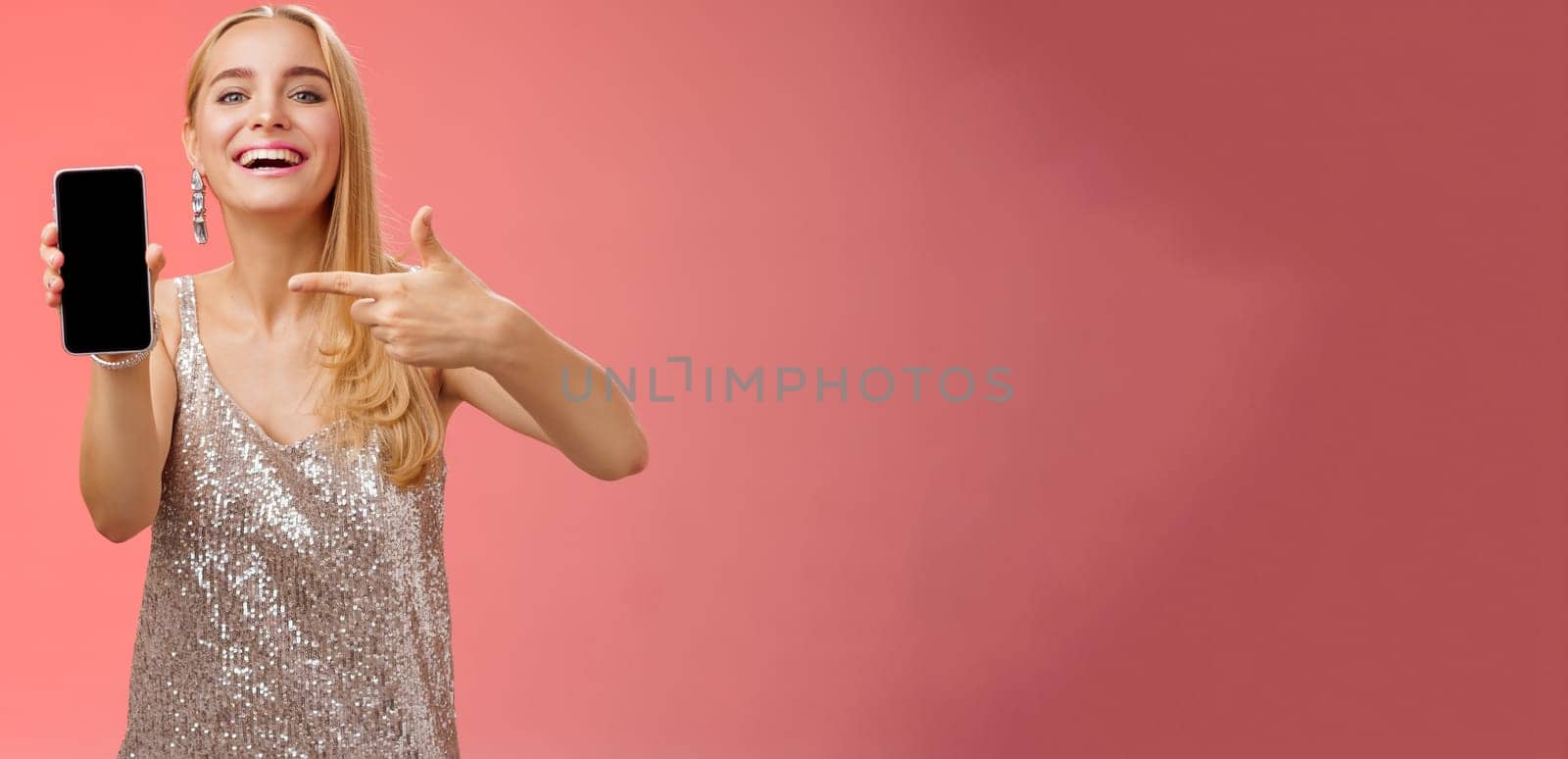 Boastful proud charming elegant blond woman in stylish evening dress show smartphone display proudly pointing mobile phone screen smiling showing photo boyfriend, standing red background by Benzoix