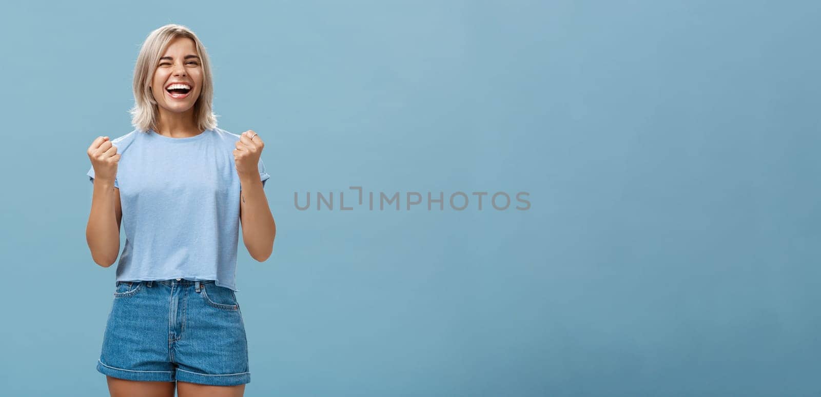 Lucky happy girl triumphing and laughing from happiness winning first place clenching fists in success gesture smiling broadly celebrating victory with enthusiasm and excitement over blue background. Emotions concept