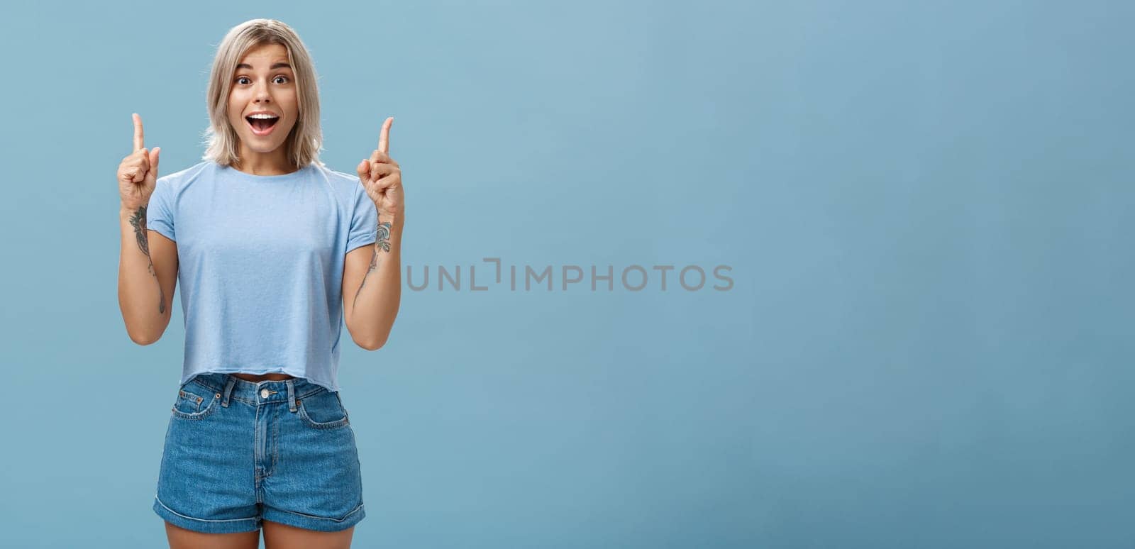 Waist-up shot of impressed enthusiastic creative blonde woman in trendy summer outfit smiling gasping being charmed and thrilled with awesome copy space pointing up over blue background by Benzoix