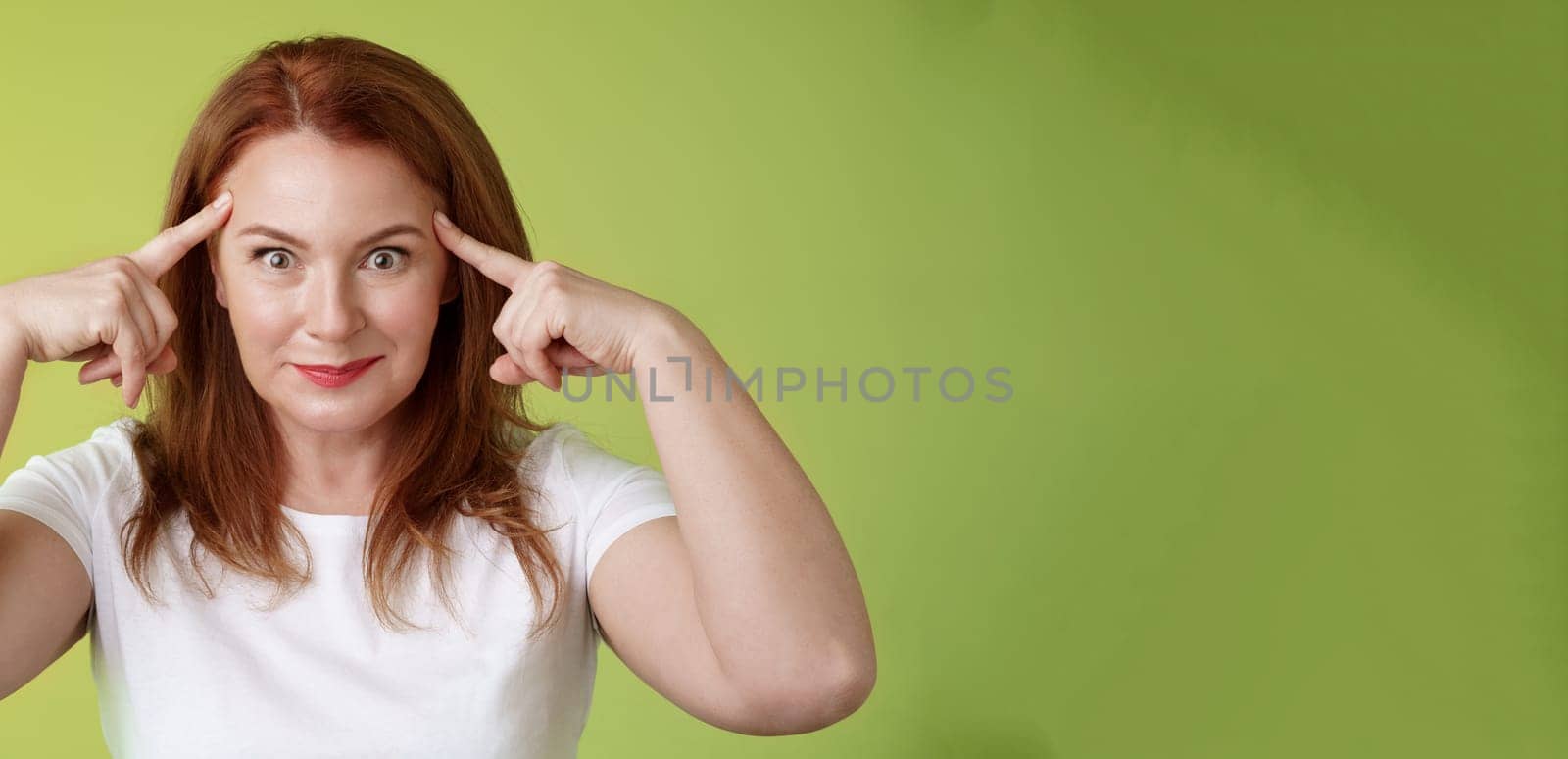 Woman playfully staring funny control your mind. Silly redhead middle-aged female touch temples popping eyes smiling delighted read thoughts trying guess intrigued what thinking green background by Benzoix