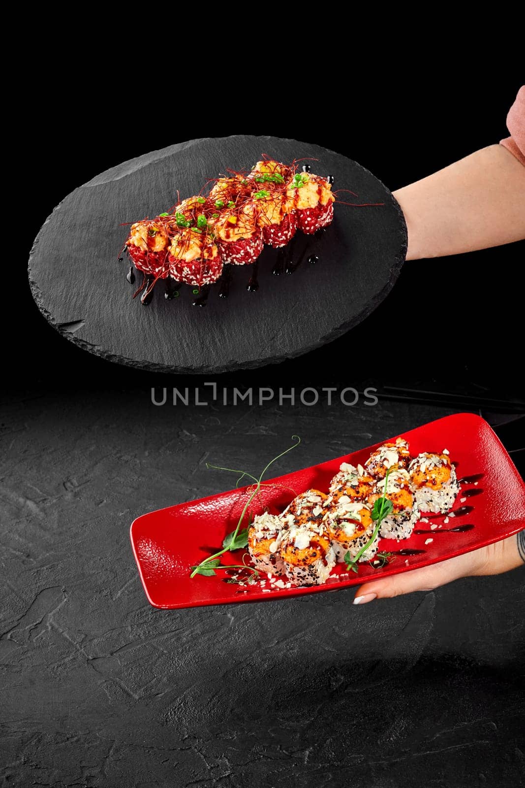 Female hands presenting two varieties of appetizing sushi rolls topped with cheese caps on sophisticated red plate and round black stone tray against dark backdrop. Japanese cuisine