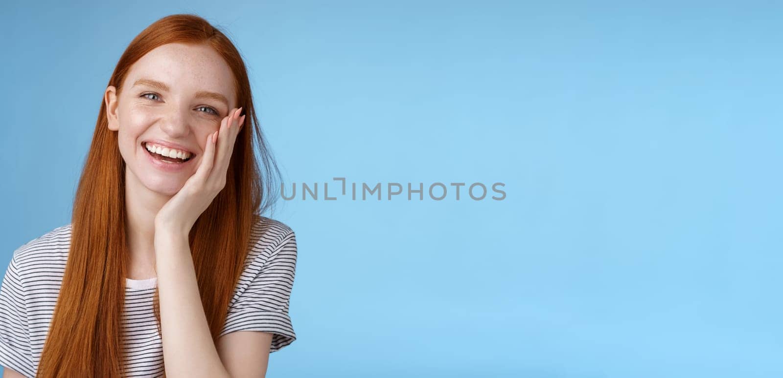 Charismatic talkative friendly-looking happy laughing redhead girl having fun discuss previous summer holidays make jokes chuckling touching face amused standing cheerful blue background.