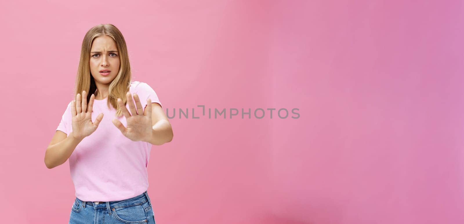 Stay away from me. Intense worried and displeased woman being victim of sexual harassment raising palms in defensive gesture making step backwards frowning asking stop, refusing over pink wall. Body language concept