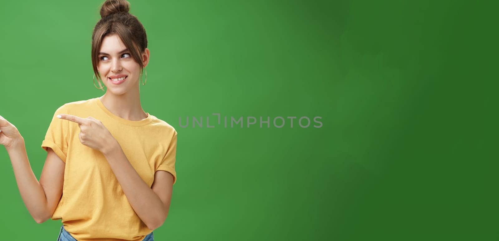 Waist-up shot of charismatic happy and carefree charming woman in yellow t-shirt pointing and looking left enthusiastic and pleased smiling cheerfully posing against green background delighted by Benzoix