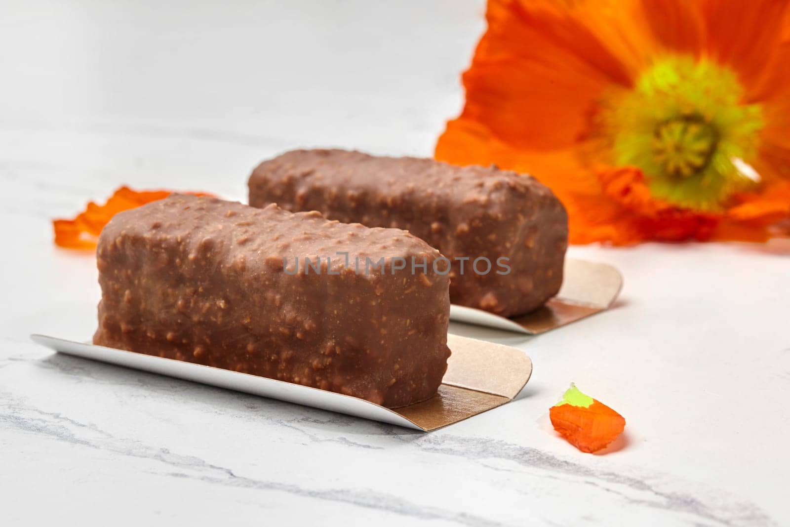 Two mini loaf cakes covered with milk chocolate and nuts, presented on marble surface with vibrant orange poppy flower in background for delectable treat