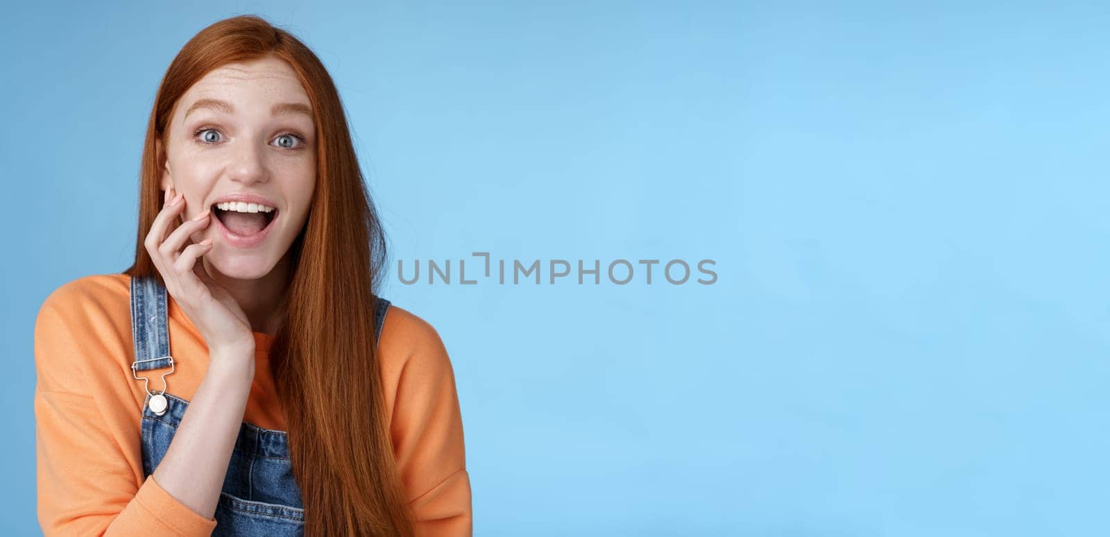 Excited enthusiasitc charismatic redhead female friend look surprised amused touch cheek open mouth thrilled discussing future prom rejoicing standing blue background joyful delighted by Benzoix