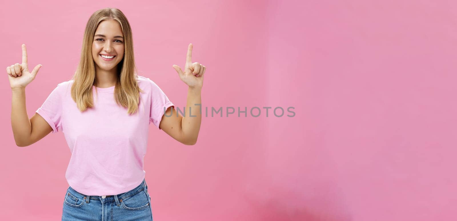 Indoor shot of pleasant attractive friendly-looking girl with tanned skin in casual t-shirt and jeans raising hands pointing up and smiling broadly at camera with satisfied look over pink background by Benzoix