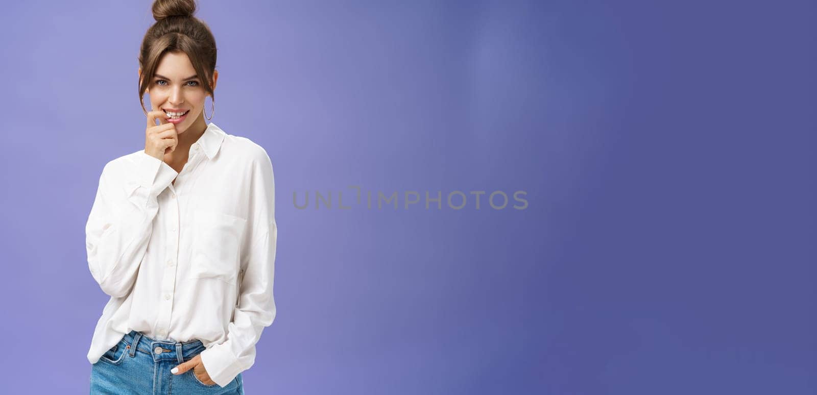 Sensual and flirty self-assured attractive woman in stylish white blouse with good self-esteem holding hand in pocket biting finger showing seducing skills looking daring at camera over purple wall. Body language concept