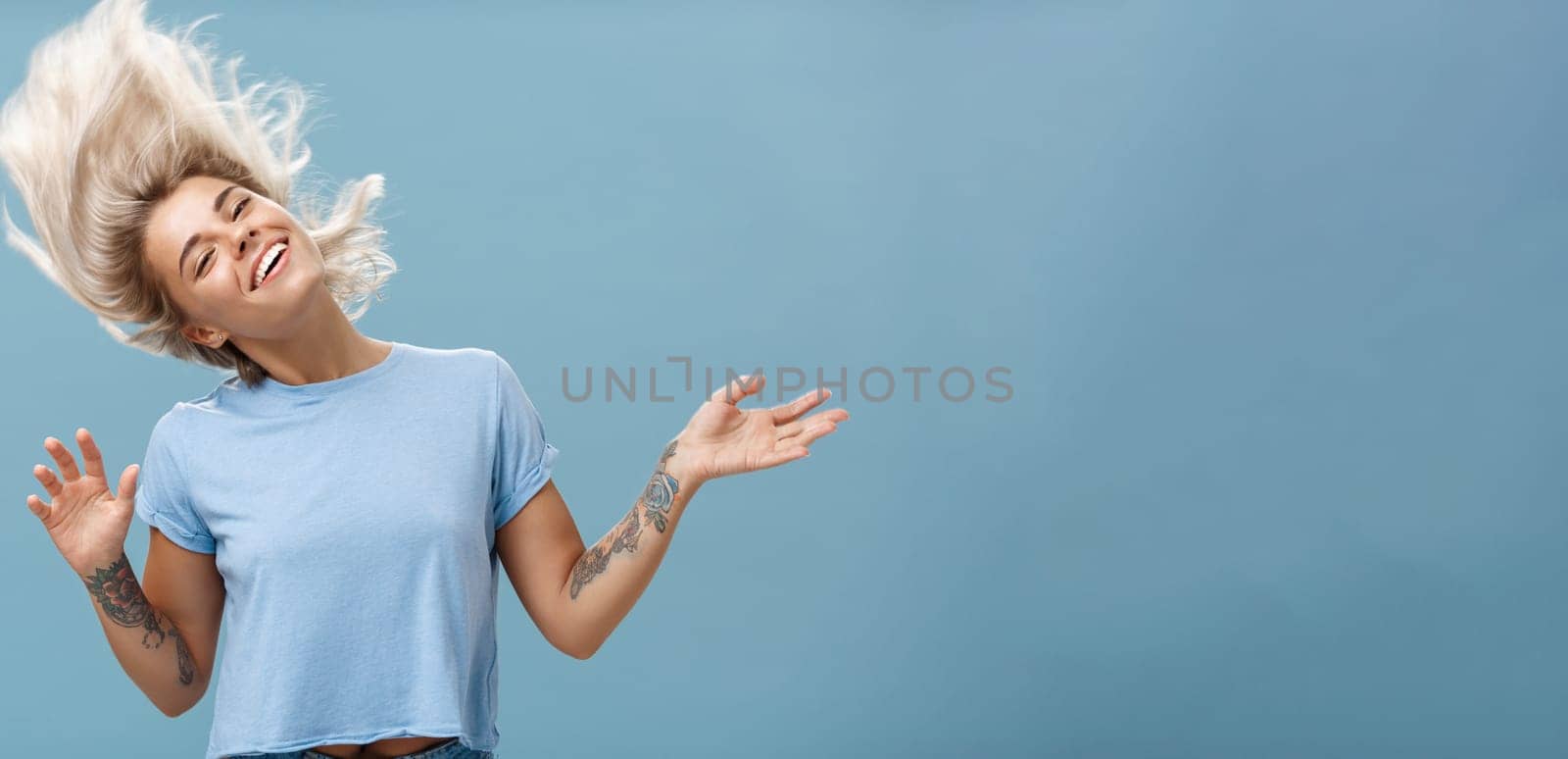 Waist-up shot of artistic and creative attractive carefree blonde female waving head and hair joyfully dancing and jumping having fun smiling broadly over blue background. Emotions and people concept