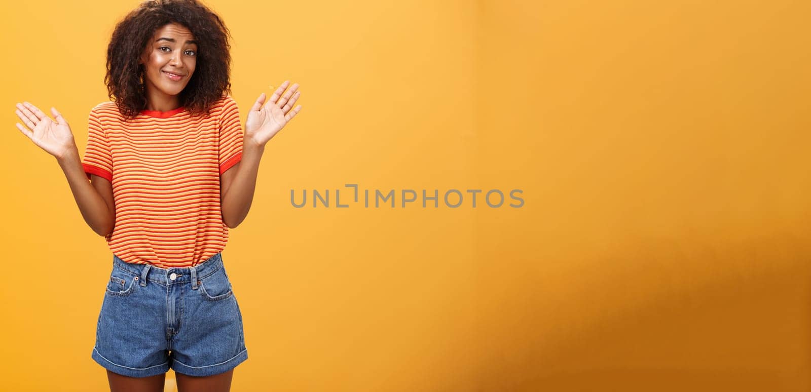 No idea do not care. Careless and indifferent calm happy african american woman with curly hair in summer clothes raising palms and shrugging in uninvolved pose smiling carefree being unaware. Body language concept