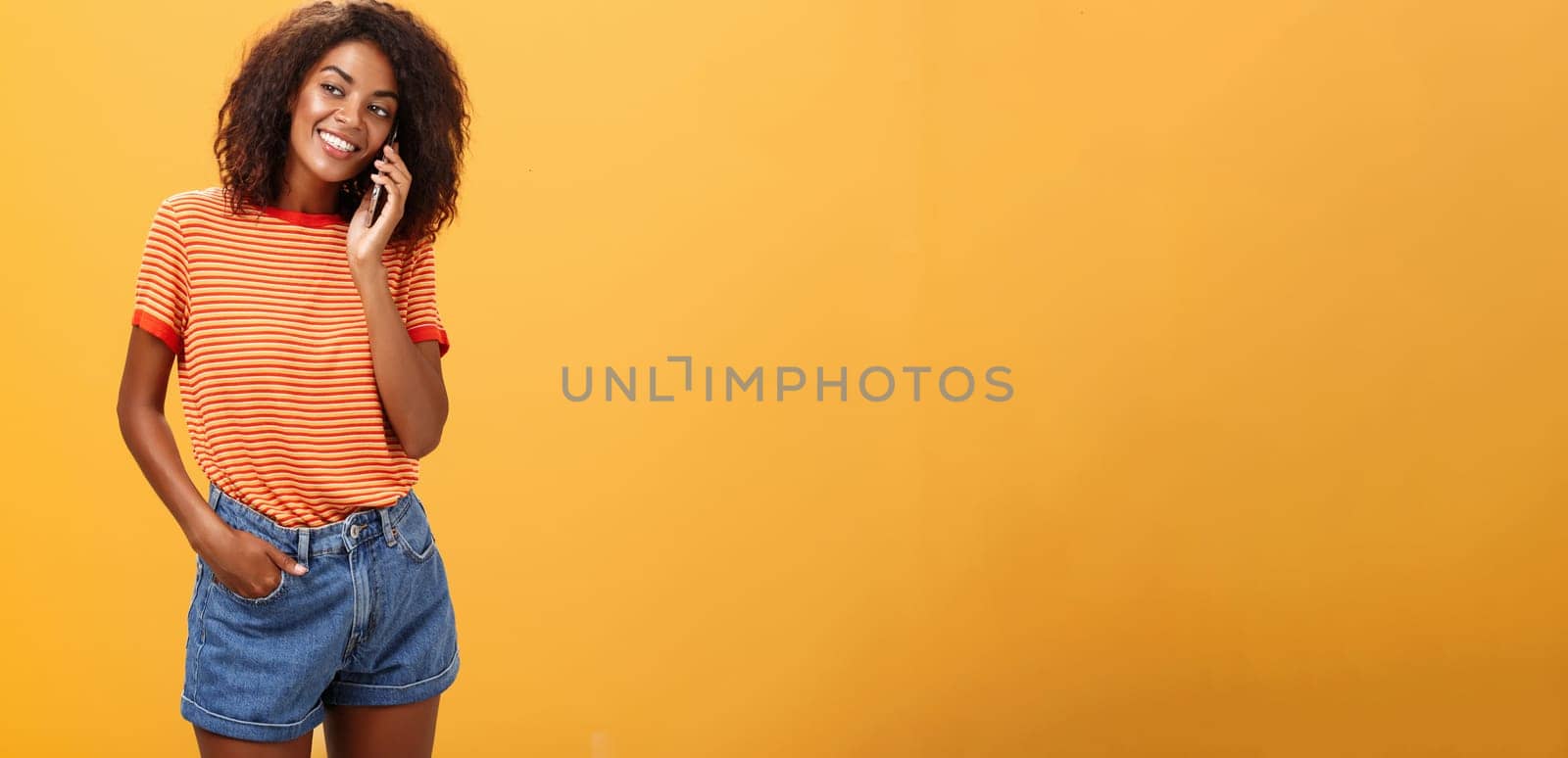 Stylish dark-skinned girl making casual phone call to friend telling all details of after romantic date standing pleased and carefree over orange background in striped t-shirt gazing left with grin by Benzoix