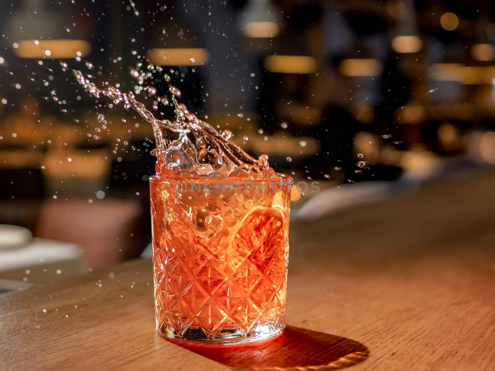 Orange or red drink with splash and droplets and orange slice garnish on bar counter. Red brown alcohol cocktail with splashes in pub or nightclub or restaurant