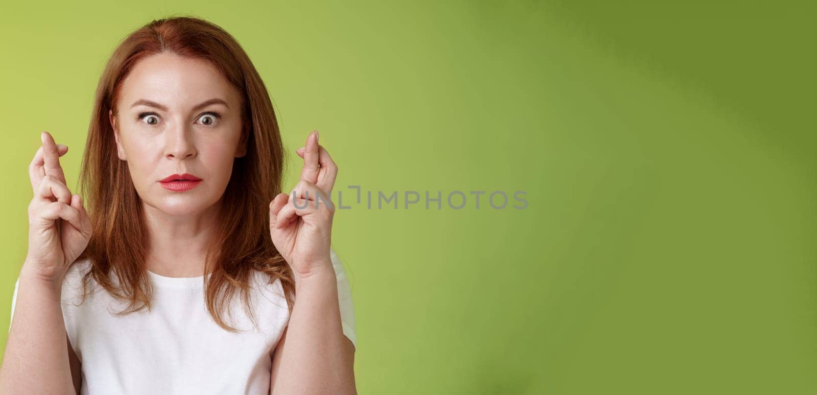 Stunned intense worried middle-aged redhead woman stare nervously awaiting important result implore dream come true receive positive reply standing stupor green background make wish good luck by Benzoix