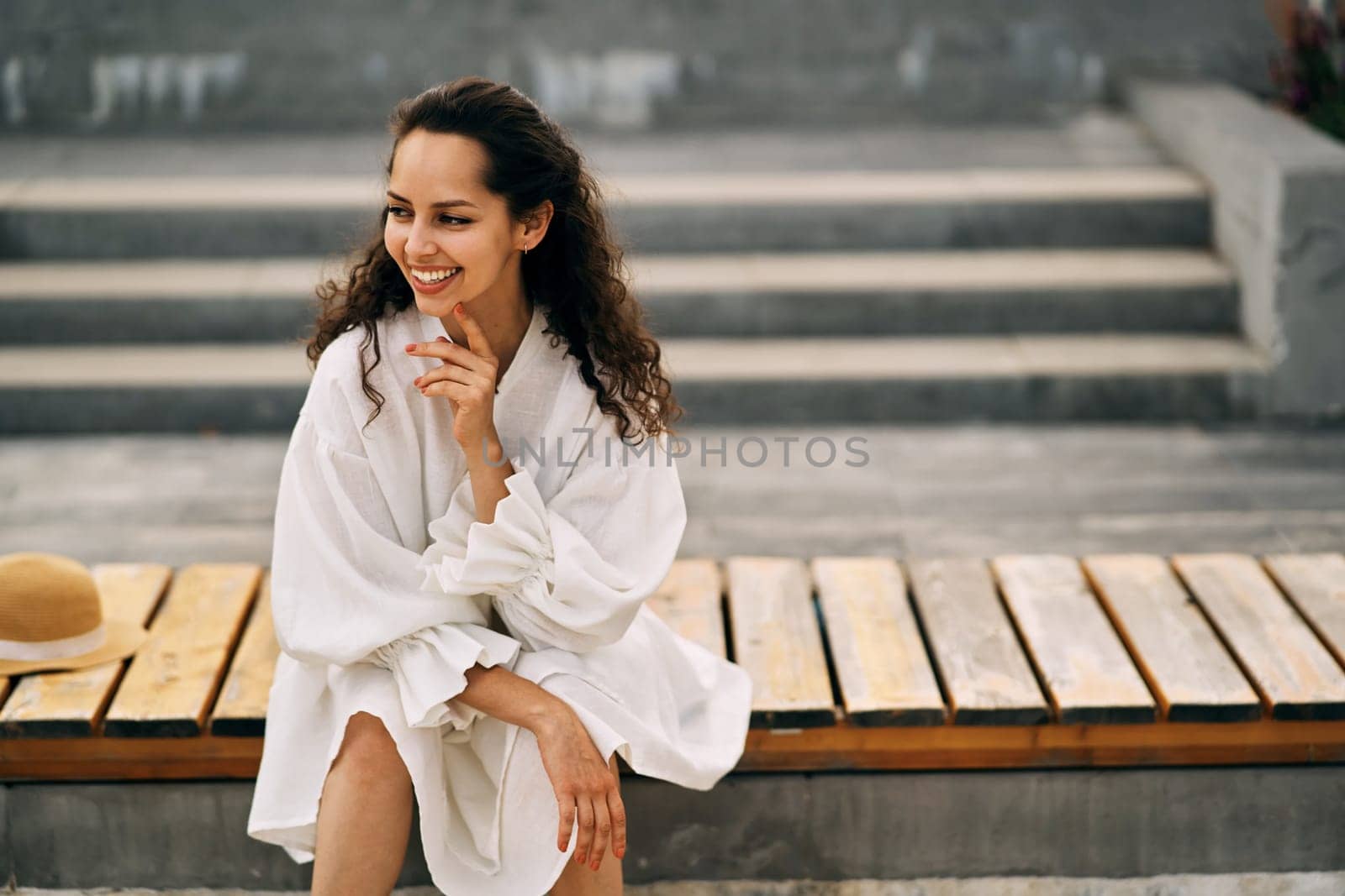 Smiling curly brunette girl in a white dress sitting on a wooden bench in the park. High quality photo