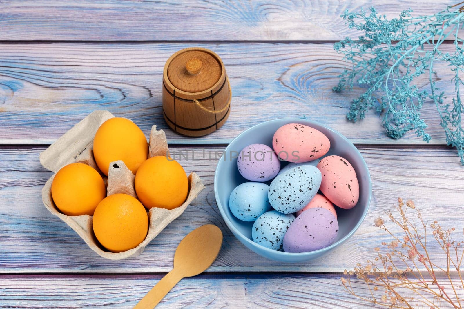 Bowl with colored Easter eggs and a cardboard box with fresh eggs, a small wooden barrel, a wooden spoon on the boards with decorative plants. Top view.