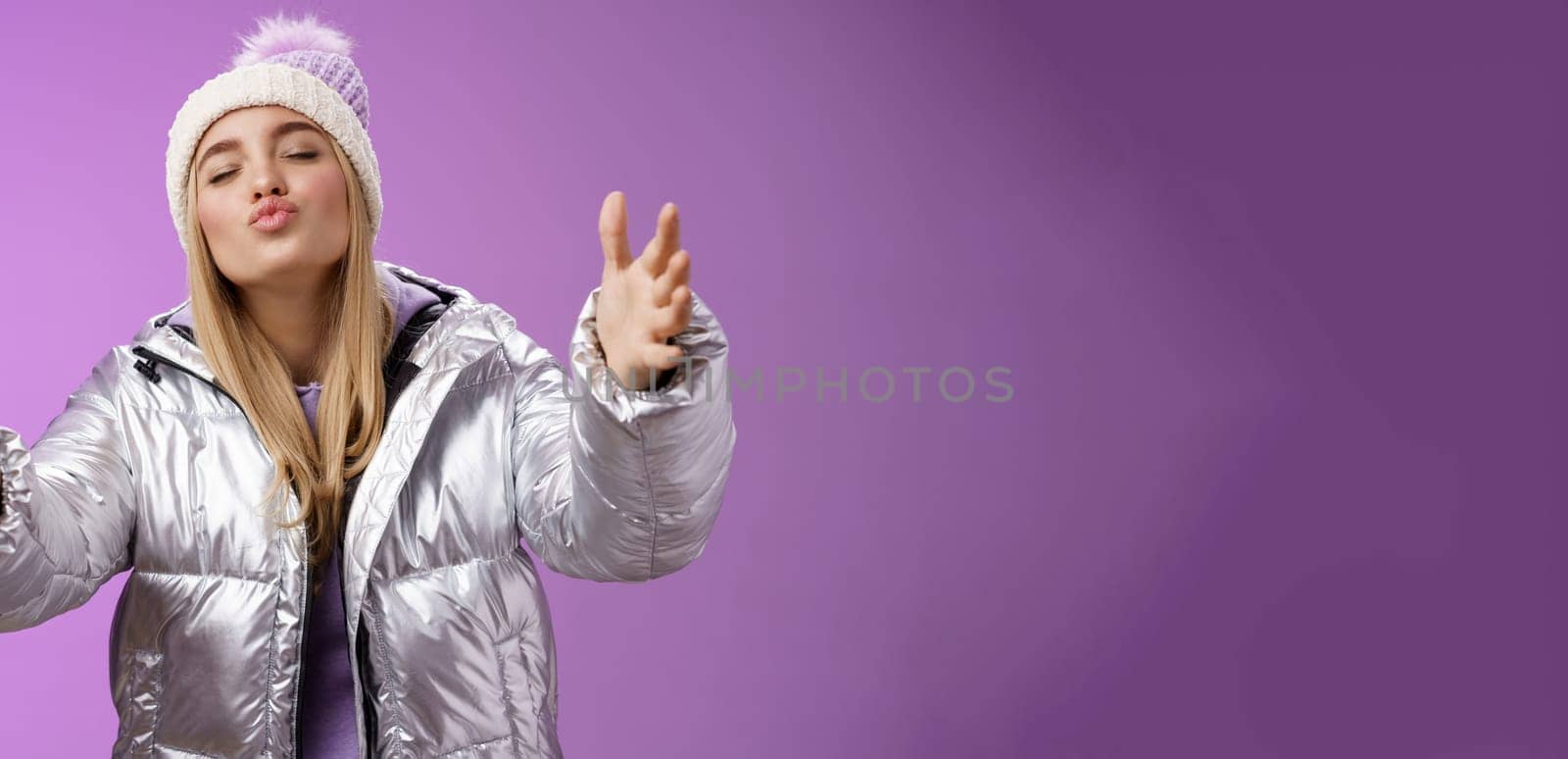 Romantic charming caucasian blond woman in cute hat silver stylish jacket pulling arms forward wanna embrace tight cuddle boyfriend folding lips close eyes give kiss, standing purple background.