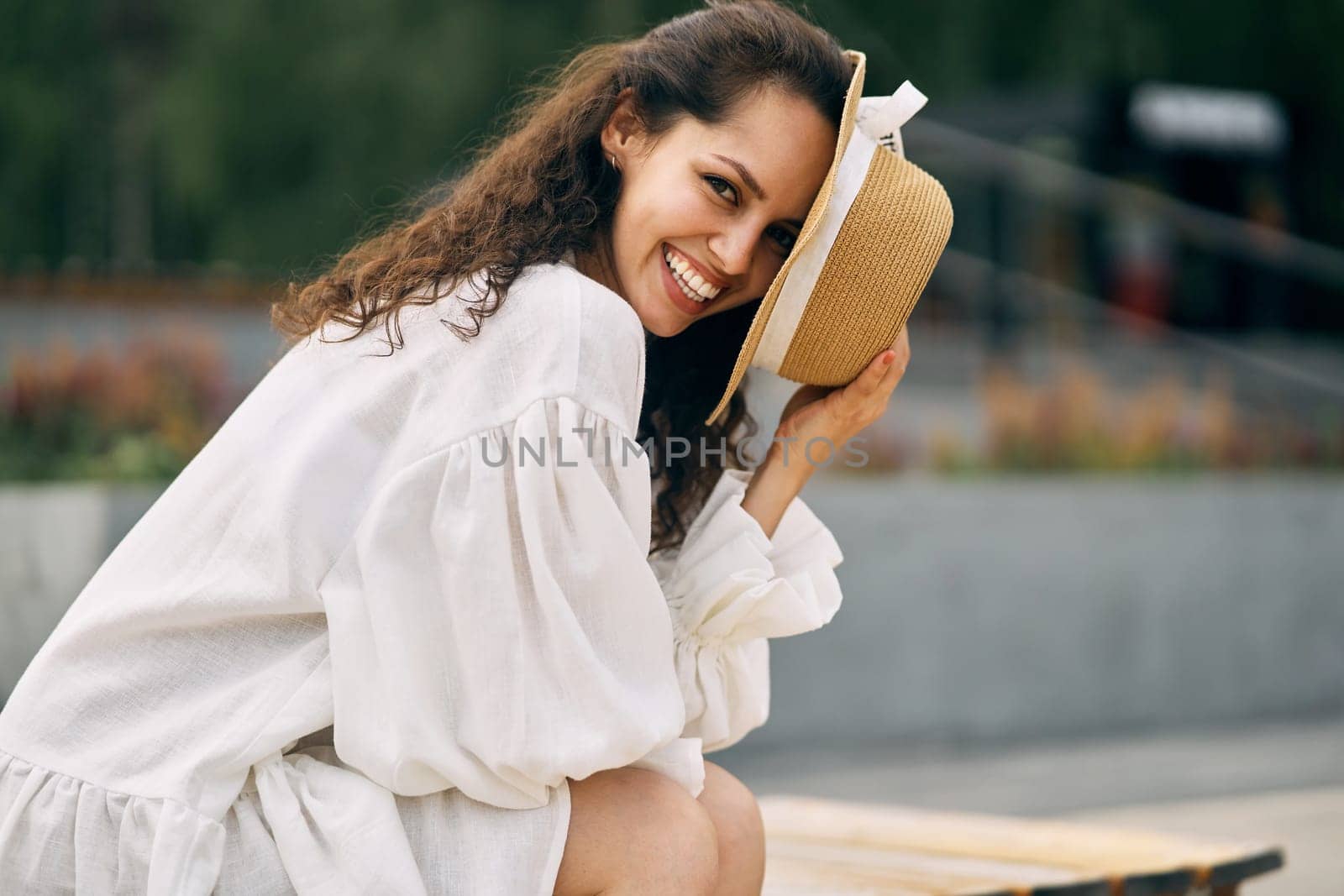 Smiling curly brunette girl in a white dress sitting and covering her face with a hat by driver-s