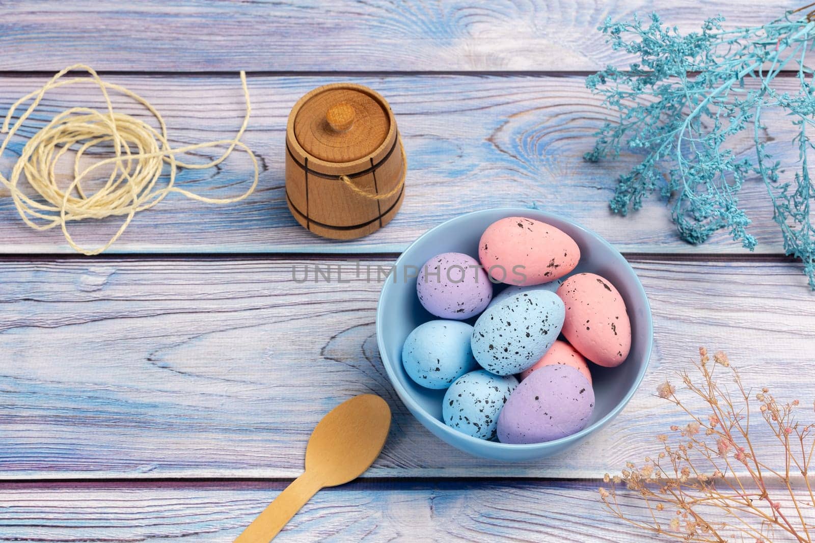 Bowl with colored Easter eggs, a small wooden barrel, a wooden spoon and a rope on the boards with decorative plants. Top view.