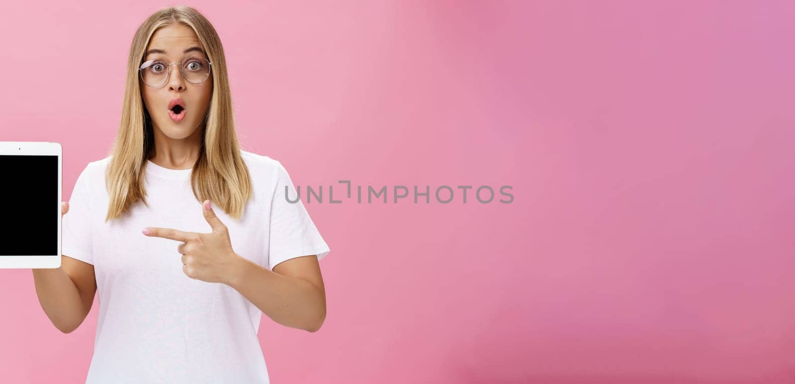 Shocked intelligent cute female in glasses and white t-shirt folding lips from amazement in wow sound gasping popping eyes amused holding digital tablet pointing at gadget screen over pink wall. Lifestyle.