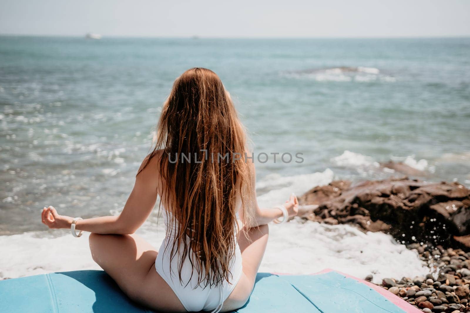 Young woman in swimsuit with long hair practicing stretching outdoors on yoga mat by the sea on a sunny day. Women's yoga fitness pilates routine. Healthy lifestyle, harmony and meditation concept.