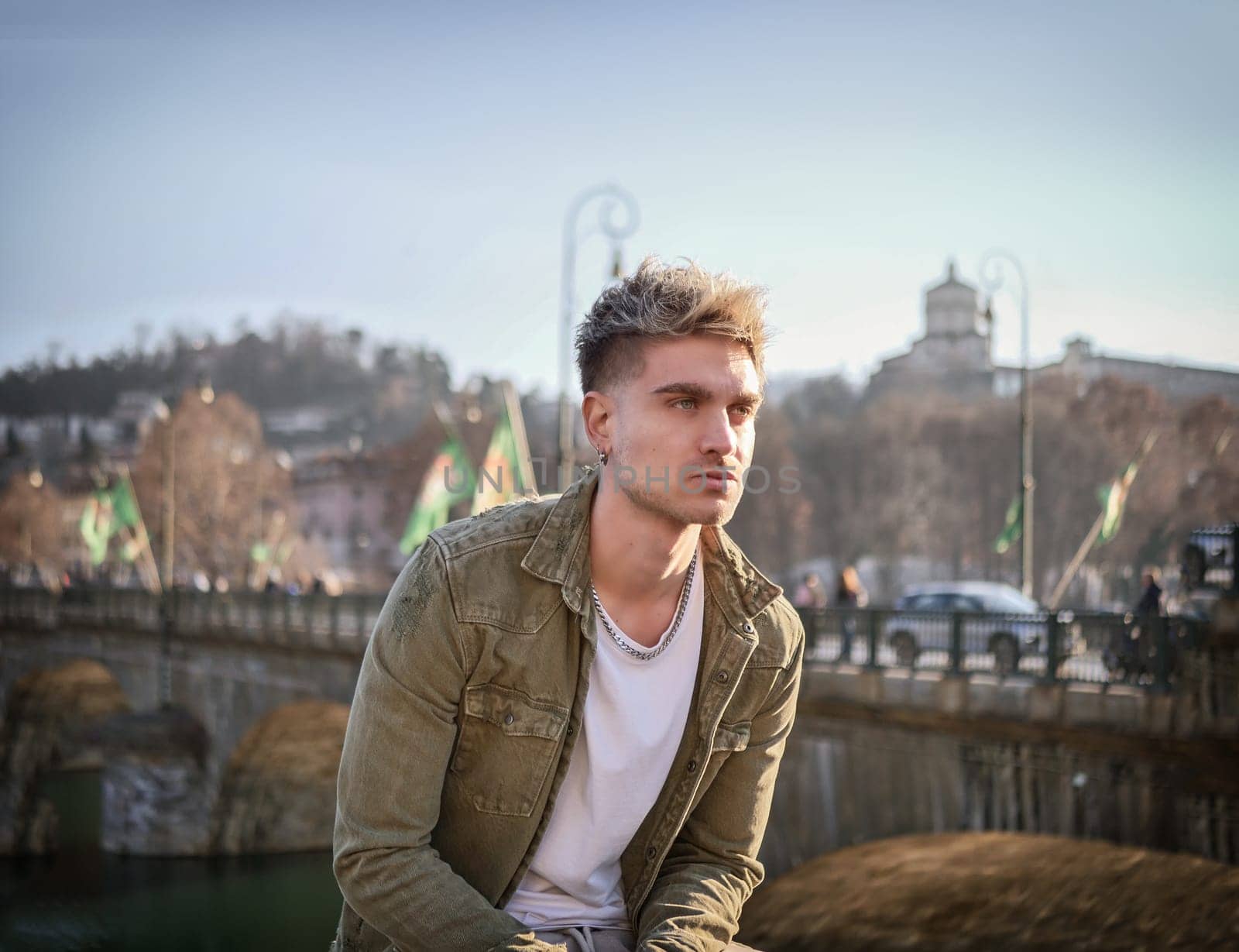 A man is seated on a ledge in front of a bridge, looking out onto the water. The scene is urban, with the bridge and surrounding buildings visible in the background.