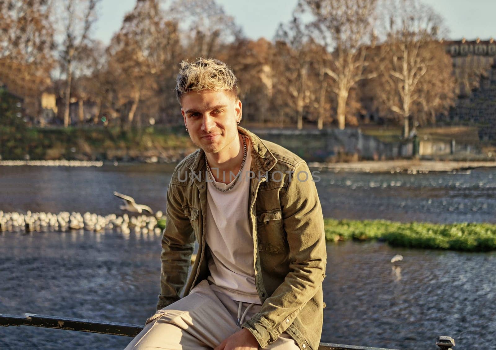 A man is seen sitting on a rail next to a body of water, gazing into the distance. The setting is serene and peaceful, with the man deep in thought.