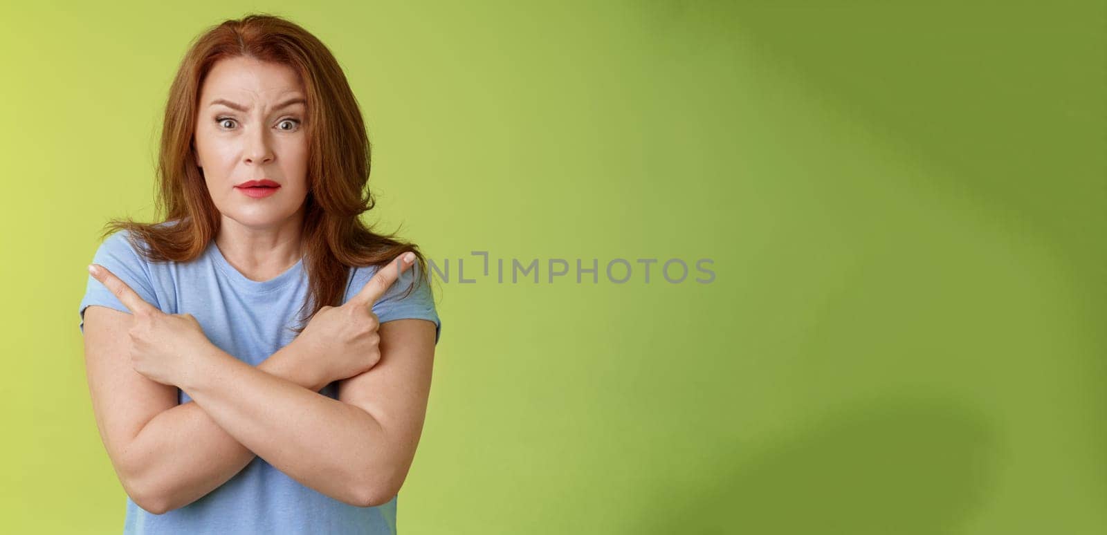 Help me choose. Worried unsure doubtful cute redhead middle-aged silly woman asking advice cross arms body pointing sideways left right look concerned camera make difficult decision green background.