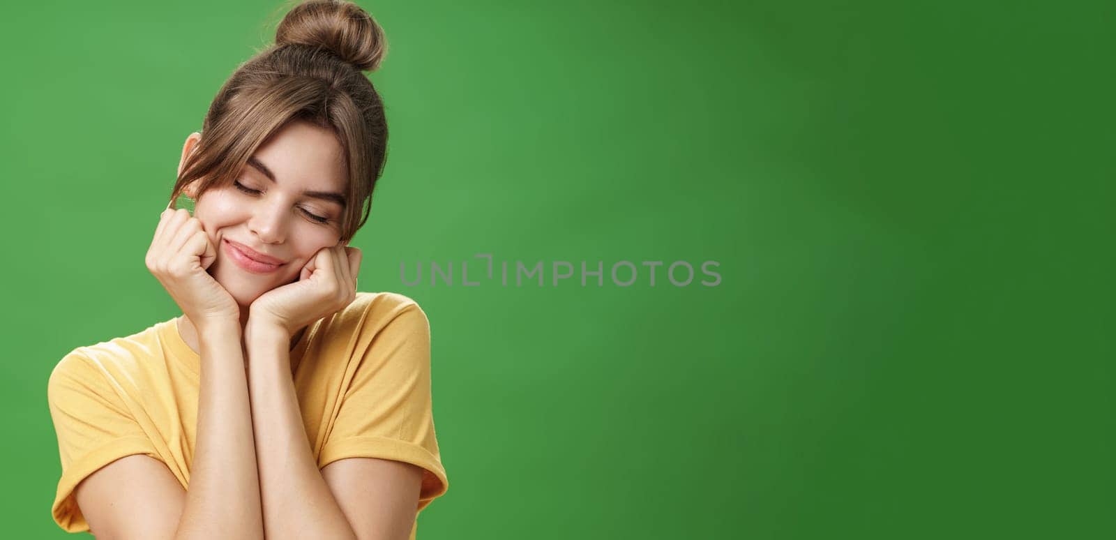 Tender sensual timid european girl in yellow t-shirt leaning head on shoulder touching cheeks with hands closing eyes and smiling with soft grin feeling nostalgic and romantic over green background by Benzoix
