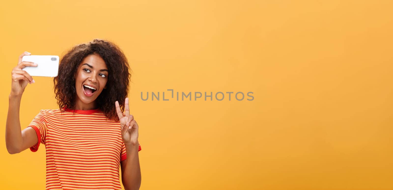 Smile to camera. Attractive and stylish self-assured dark-skinned female model with curly hairstyle showing peace gesture while taking selfie holding smartphone near face and smiling at gadget screen.