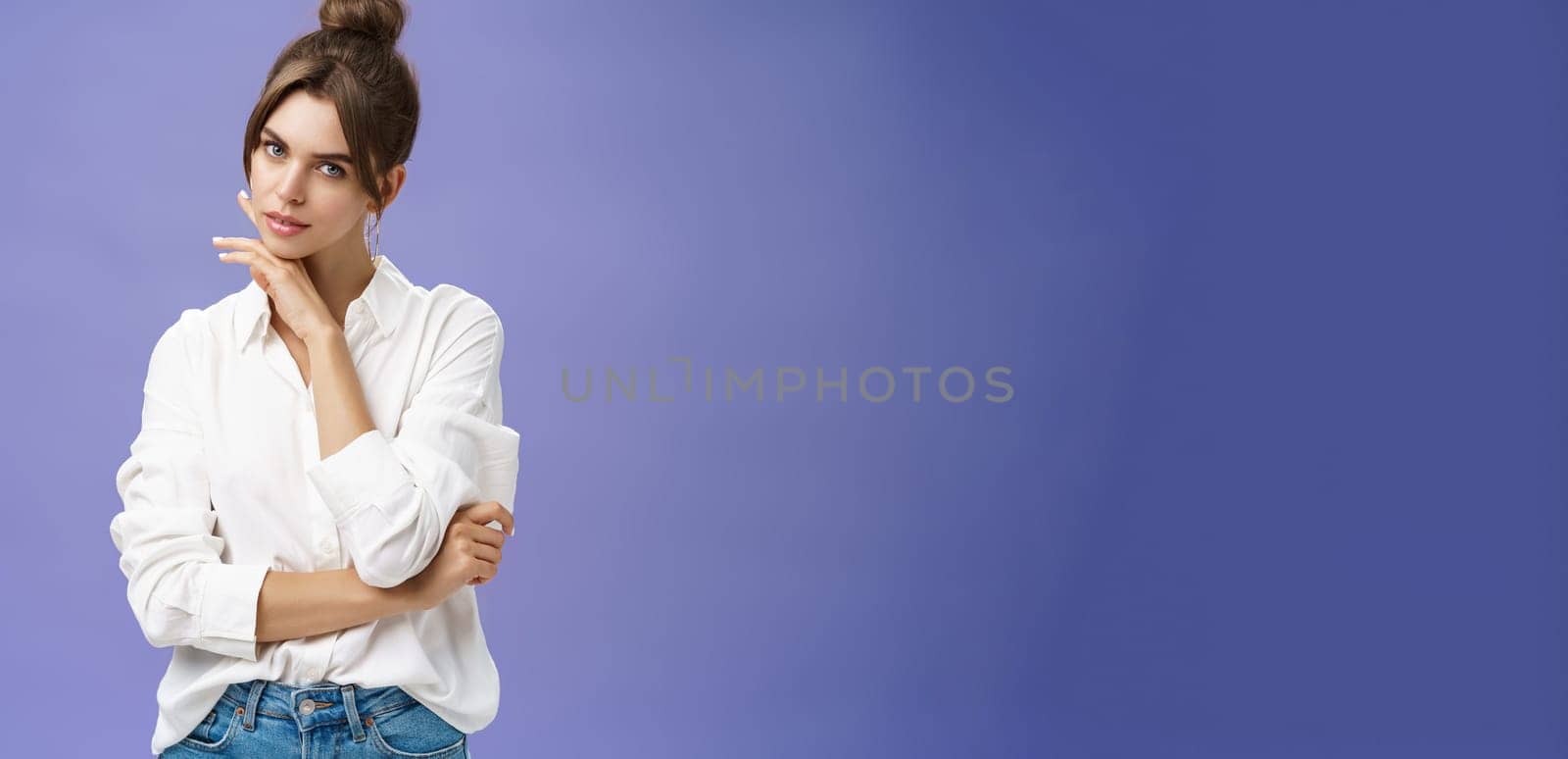 Portrait of tender and feminine stylish woman in white blouse posing sensually and flirty touching chin gazing daring at camera posing against purple background with self-assured expression by Benzoix