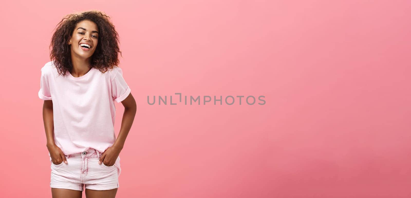 Brighten up life with smile. Carefree charismatic happy african american female with afro hairstyle in trendy outfit laughing out loud from joy and amusement holding hands in pockets over pink wall by Benzoix